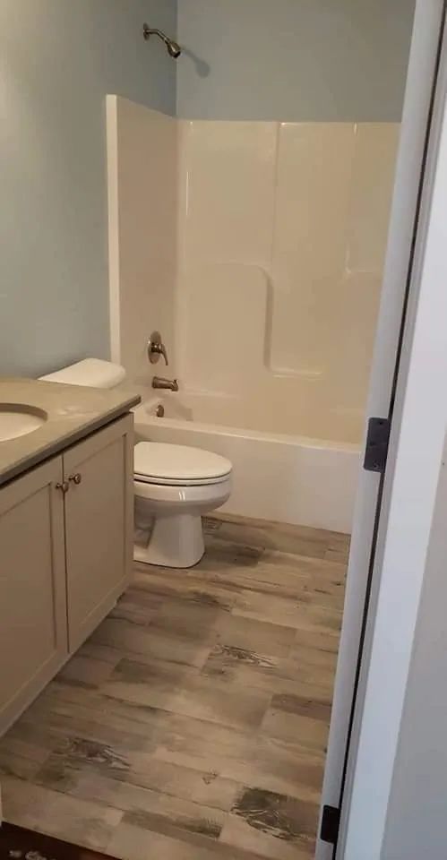 Small bathroom with toilet, bathtub, vanity sink, and wood-look flooring; shower curtain hangs at right.