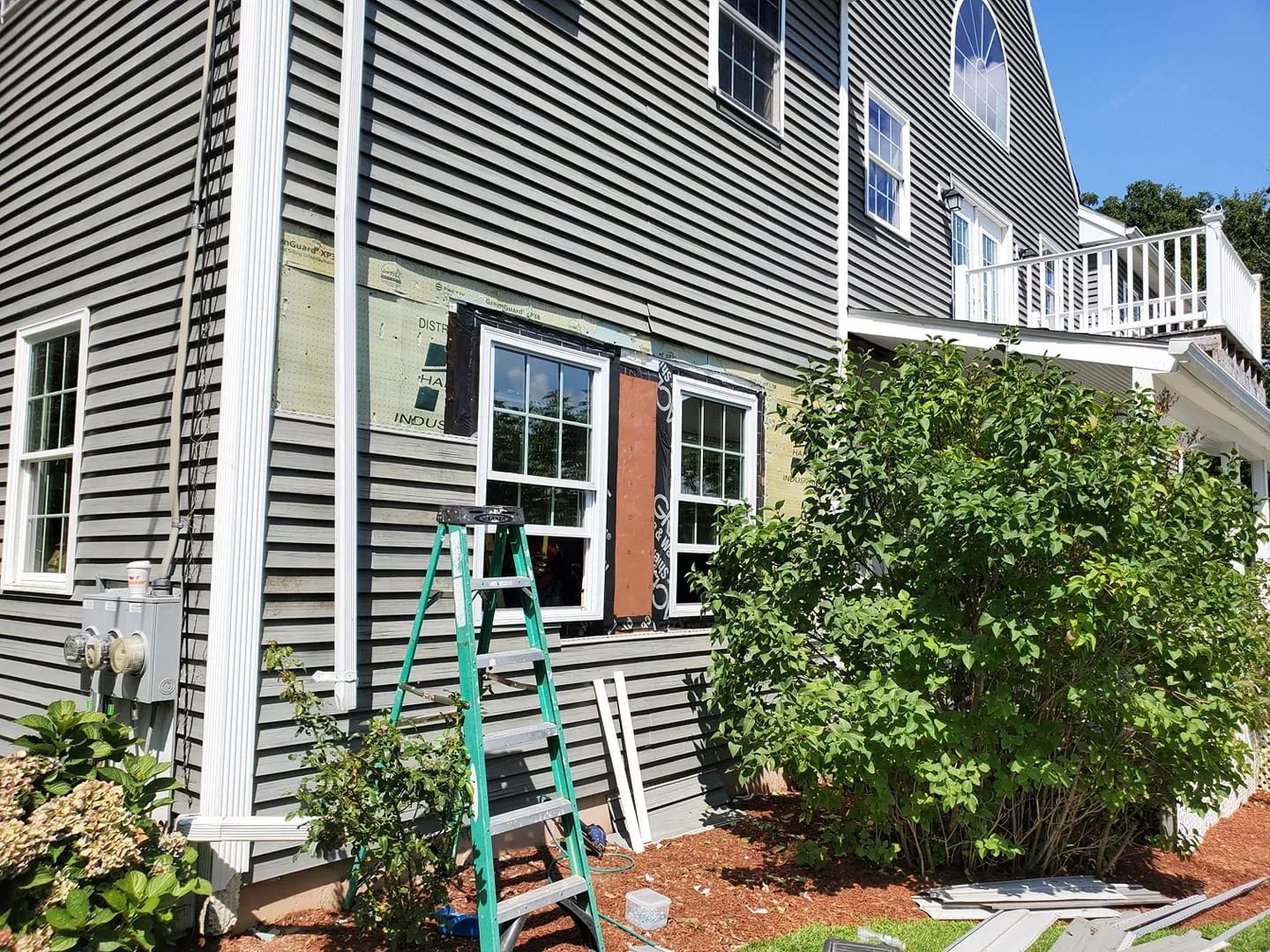Gray house exterior with a green ladder beside a white-trimmed window and bushes in front