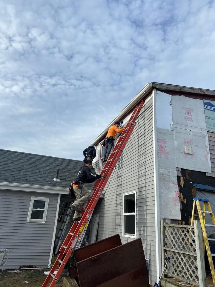 Two workers on a red ladder repairing a house roof beside white siding under a cloudy sky