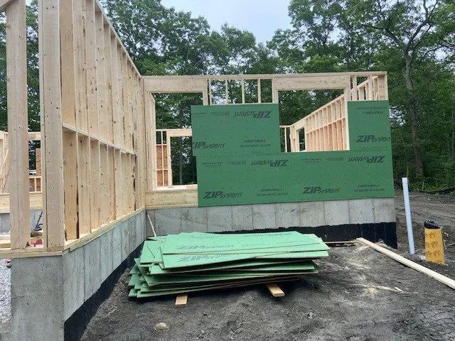 Wood-framed house under construction with green sheathing, concrete foundation, and stacked panels on the floor