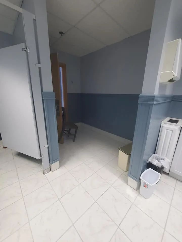Narrow hospital hallway with blue-gray walls, white tile floor, chair, and a paper towel dispenser.