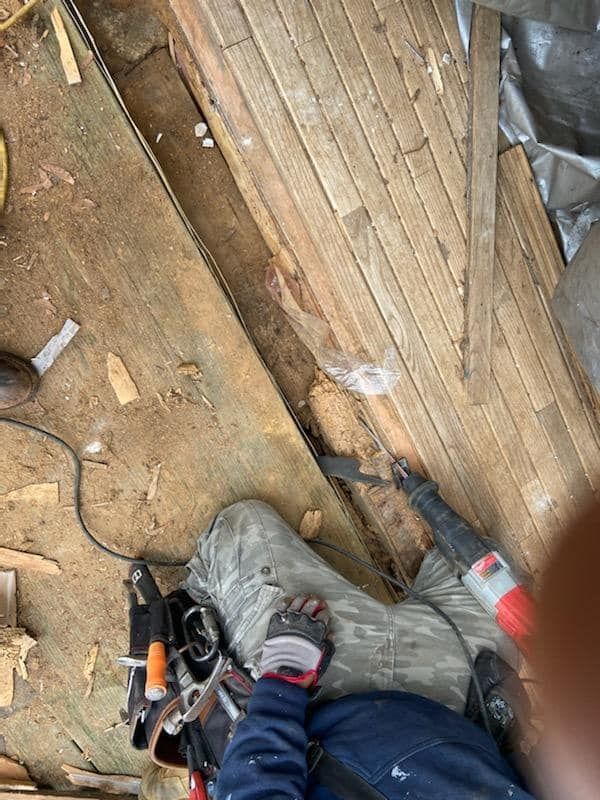 Person’s feet beside tools on a dusty wooden floor with a black cable and plastic bag.