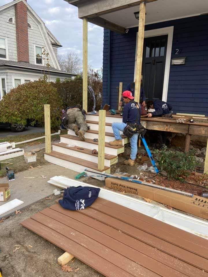 Workers building a front porch and steps on a blue house, with lumber and tools in the yard.