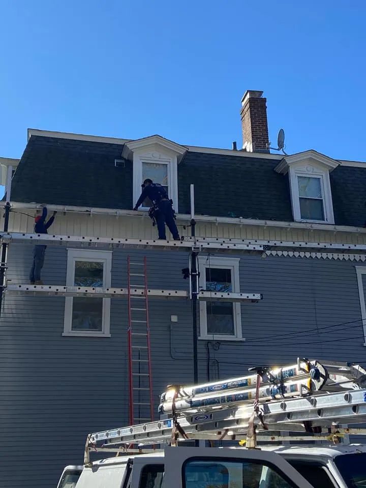 Worker on ladder repairing a gray house roof with ladders and tools under a blue sky