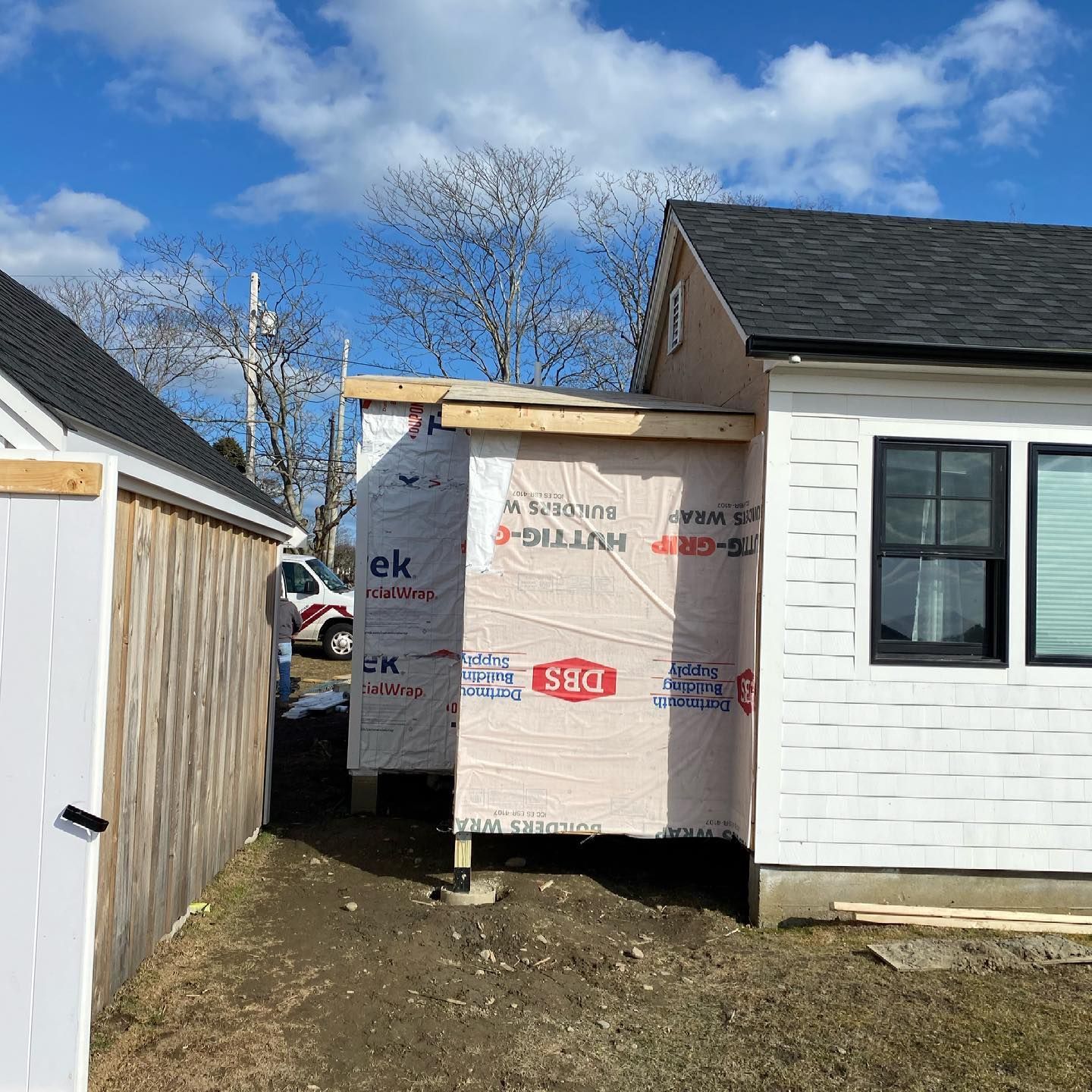 House under construction with exposed insulation beside a white house and fence on a sunny day
