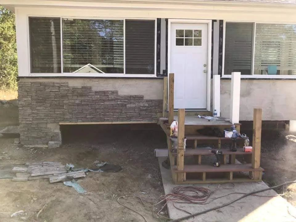 House front with damaged lower wall and unfinished wooden steps leading to a white door.