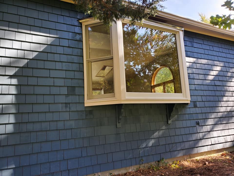 Blue shingled house wall with an open bay window and trees reflected in the glass