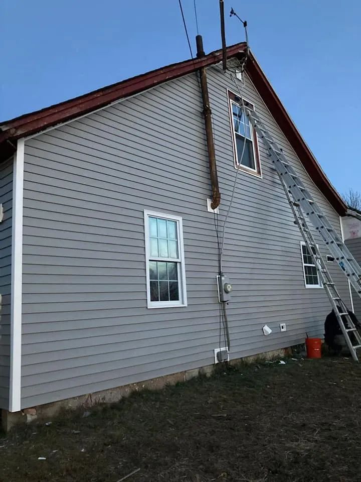 Side view of a gray house with white trim, steep roof, and an exterior ladder under a cloudy sky