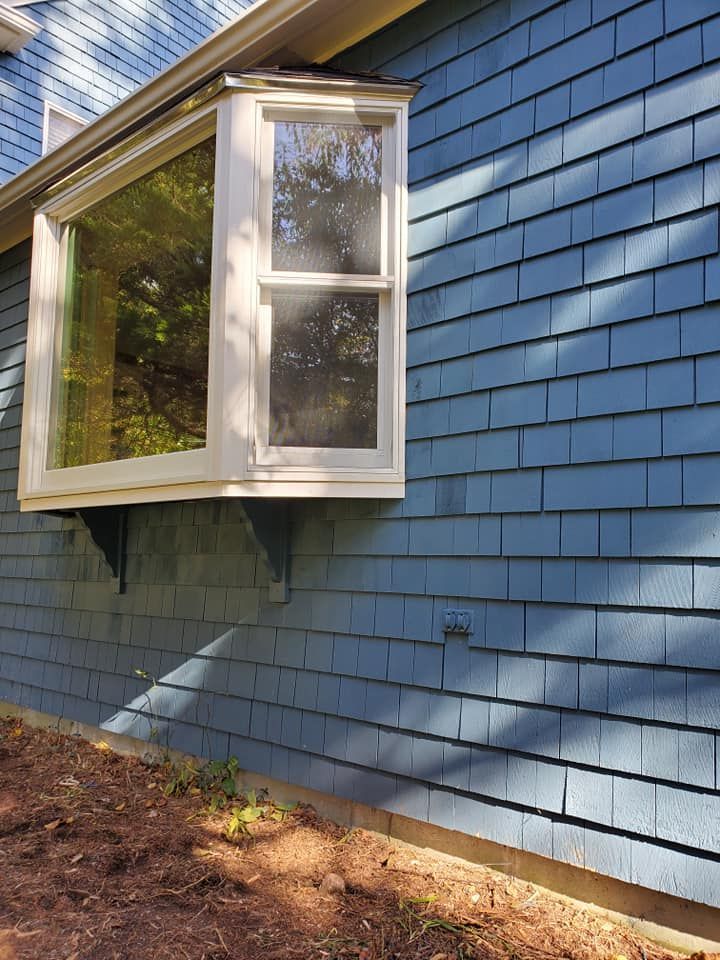 Blue house siding with a white bay window extending outward above a dirt and leaf-covered yard