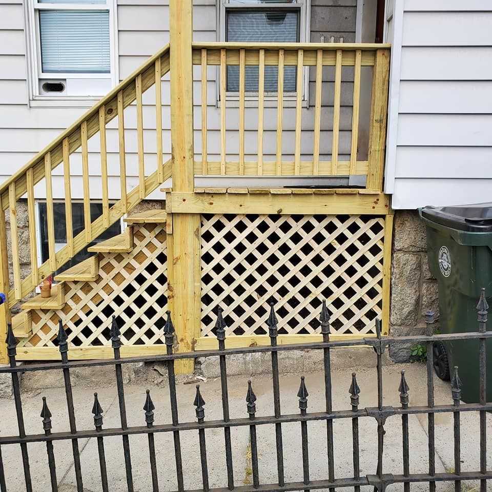 Yellow wooden porch stairs and lattice skirting on a house beside green trash bins and a black fence.