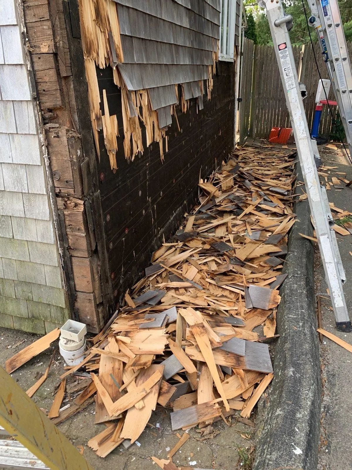 Damaged exterior wall with missing wood shingles and debris piled along a narrow walkway beside a ladder