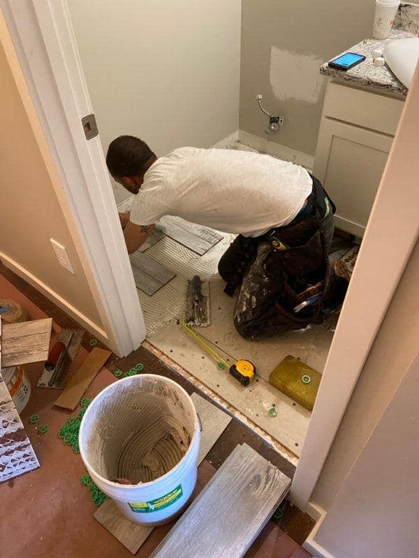 Person kneeling to work under a bathroom sink amid tools, debris, and a bucket on the floor.