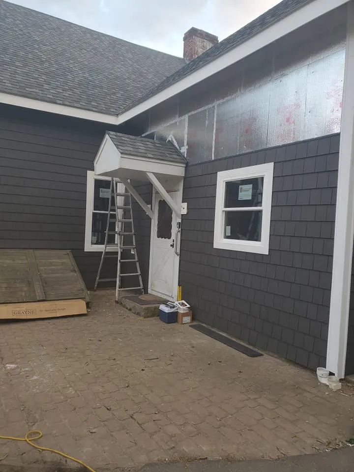 Exterior of a house with a small covered entry, ladder, and stacked boards on a paved patio, during renovation