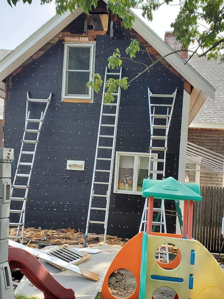 House exterior with dark blue siding and three ladders leaning against it, under construction in a yard
