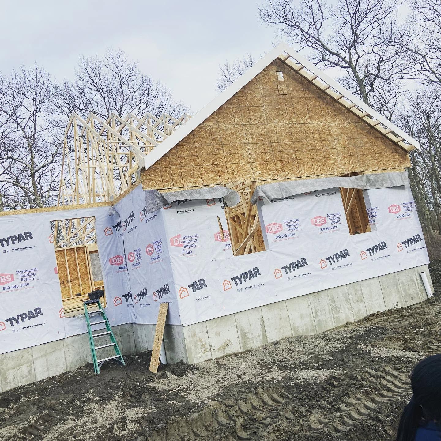 Partially built house with white Typar wrap, exposed brick gable, and a green ladder on muddy ground