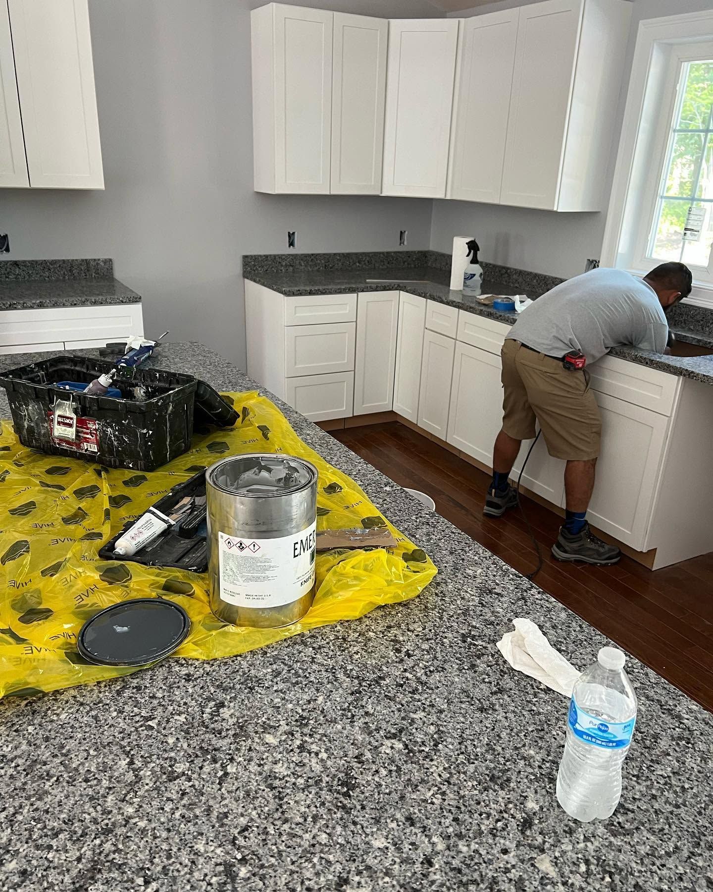 Man painting white kitchen cabinets beside a black granite counter and paint supplies on the floor.