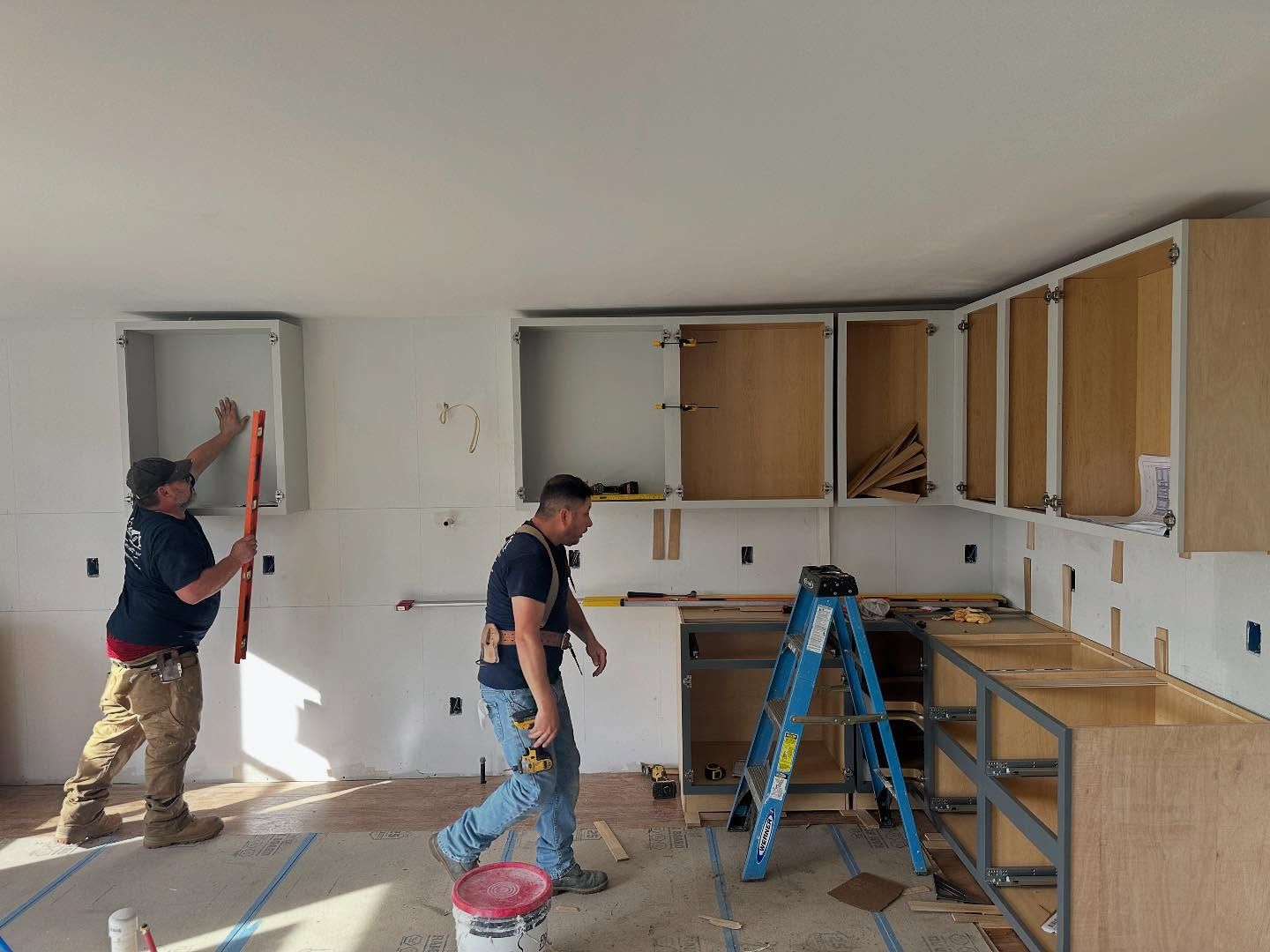 Workers renovating a kitchen with white cabinets, blue ladder, and tools scattered on the floor.
