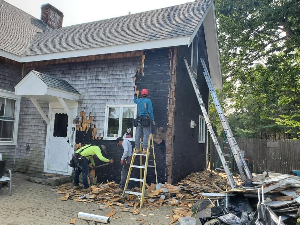 Workers repairing house exterior with ladders, exposed siding, and debris on the ground