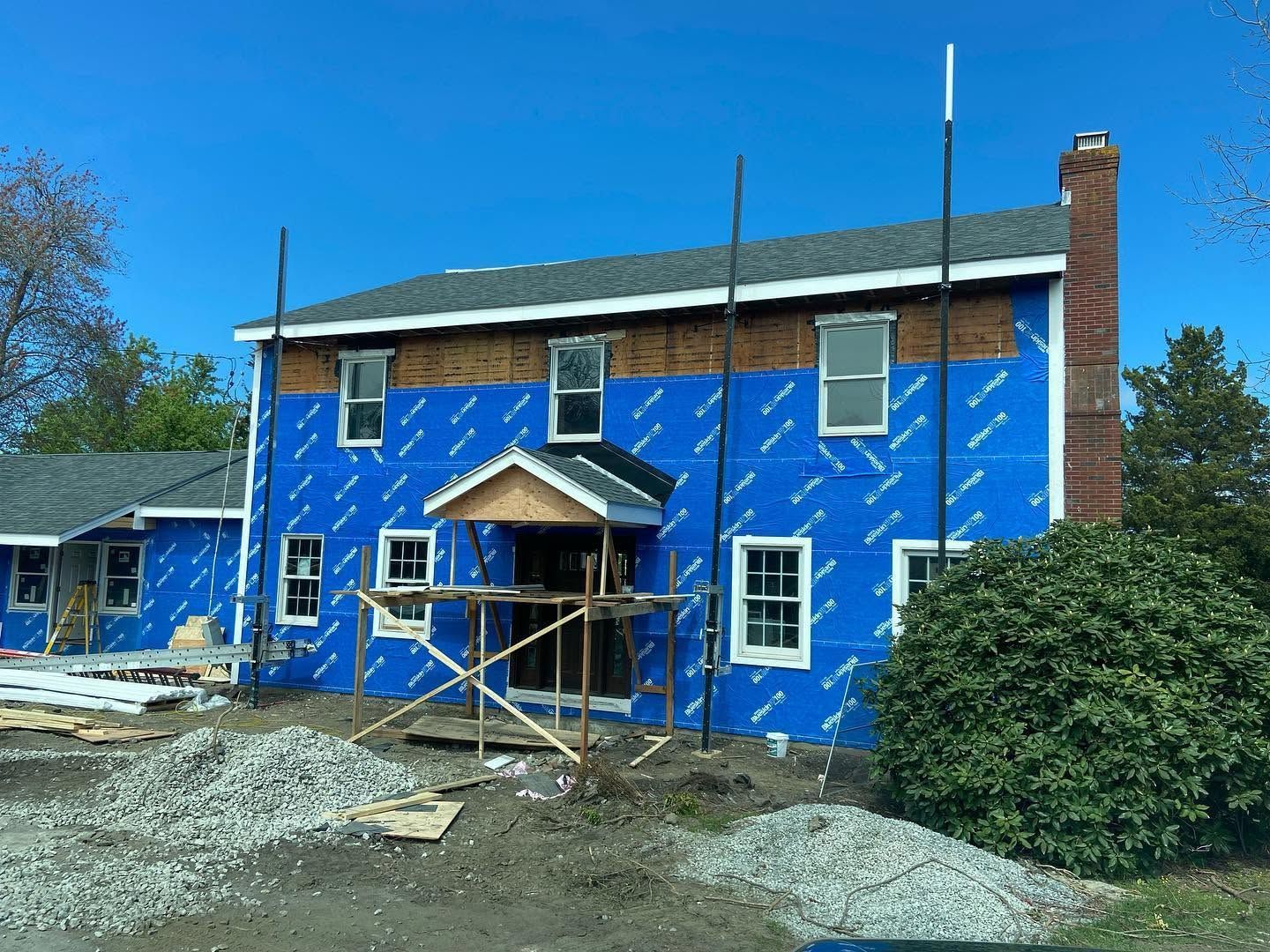 Blue house under construction with scaffolding, white trim, and a brick chimney on a gravel lot.