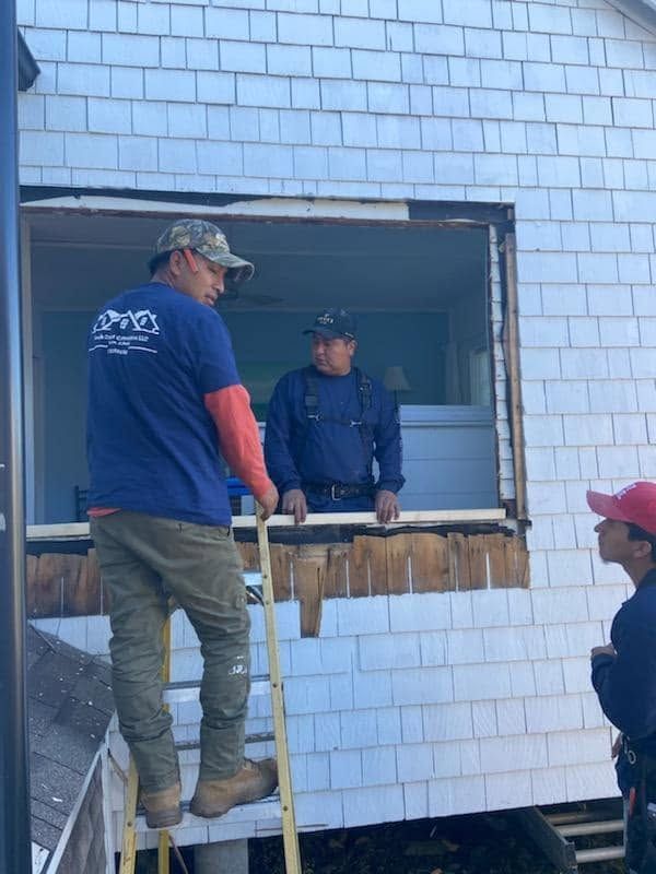 Two workers at a broken window on a white house exterior, one standing on a ledge and one inside