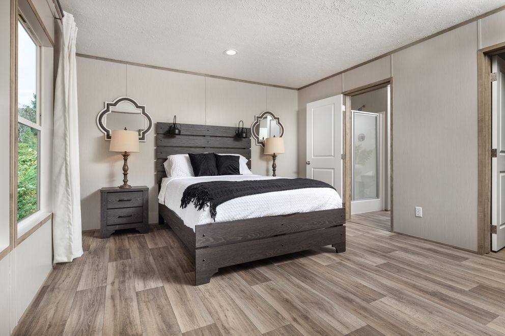 Bedroom with wooden flooring, gray walls, bed, lamps, mirrors, and an open doorway to a bathroom.