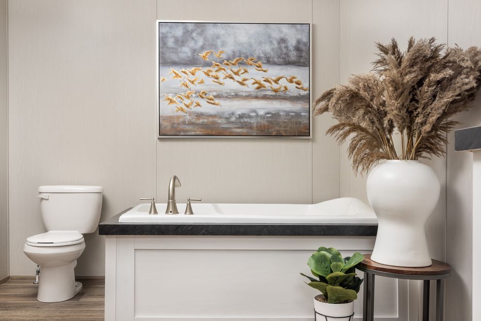 Bathroom with a white tub, toilet, and vase with dried plants. Painting and potted plant in view.