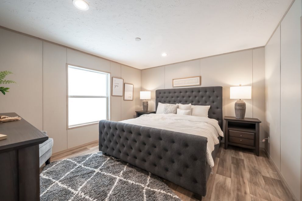 Bedroom with a large gray tufted bed, nightstands, rug, and a window with artwork.