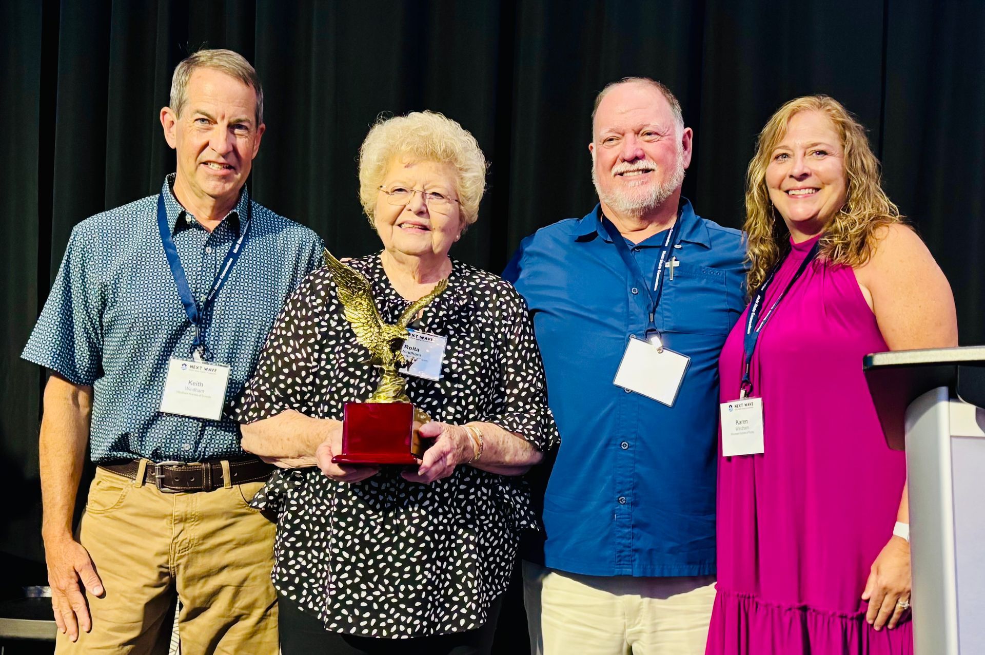 Four people stand, smiling. Woman holds golden eagle trophy. The setting is indoors, stage with curtains.