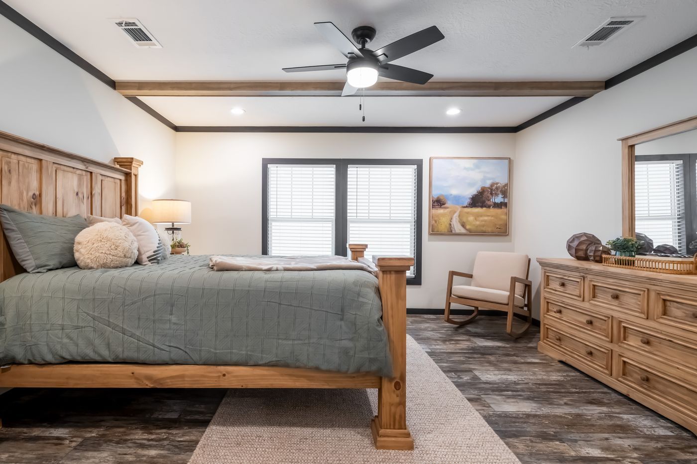 Rustic bedroom with wooden bed, dresser, and ceiling beams, with gray walls and a light blue bedspread.