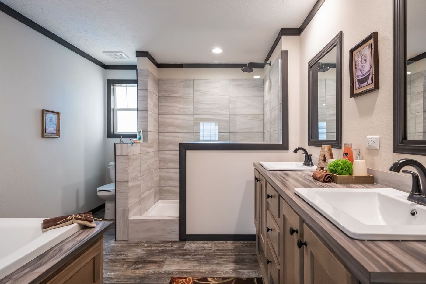 Bathroom with dual sinks, shower, and toilet. Light gray walls, wood-look floor, dark trim, and framed mirrors.