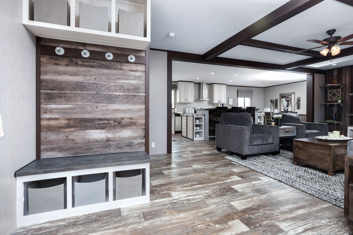 Entryway with bench and storage, leading to a living area with gray couches and a kitchen.