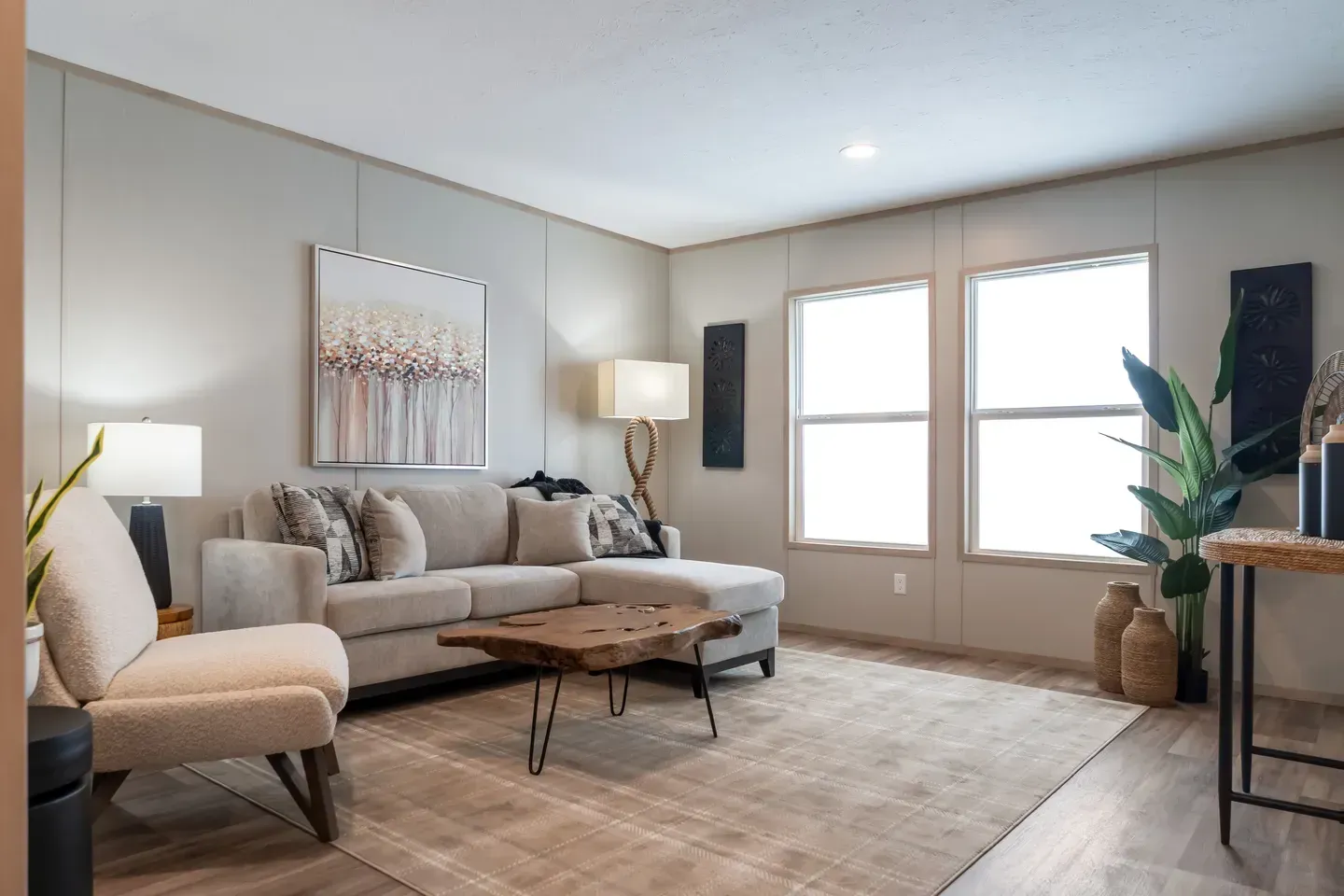 Living room with light-colored walls, neutral furniture, large windows, and a decorative plant.