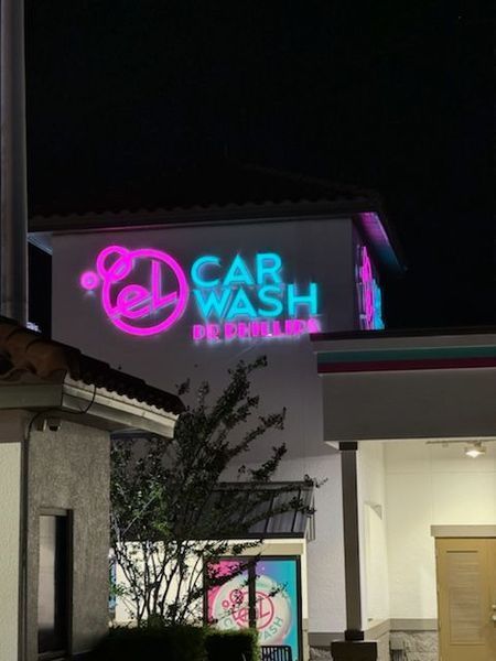 Car wash sign at night. Bright pink and blue neon text and logo above a building with some foliage.