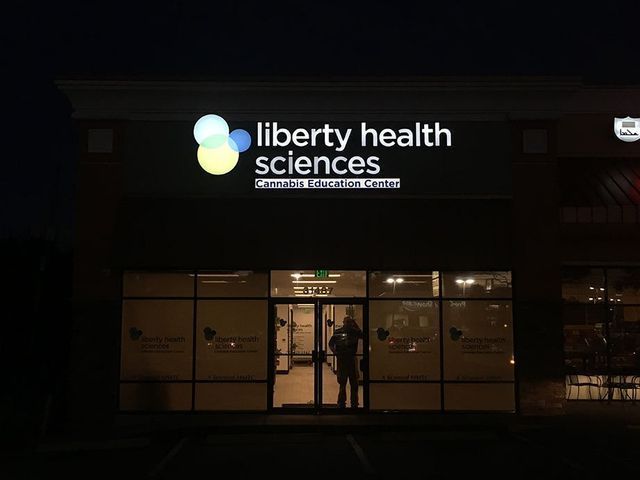 Liberty Health Sciences Cannabis Education Centre storefront at night. Illuminated sign, glass doors, person inside.