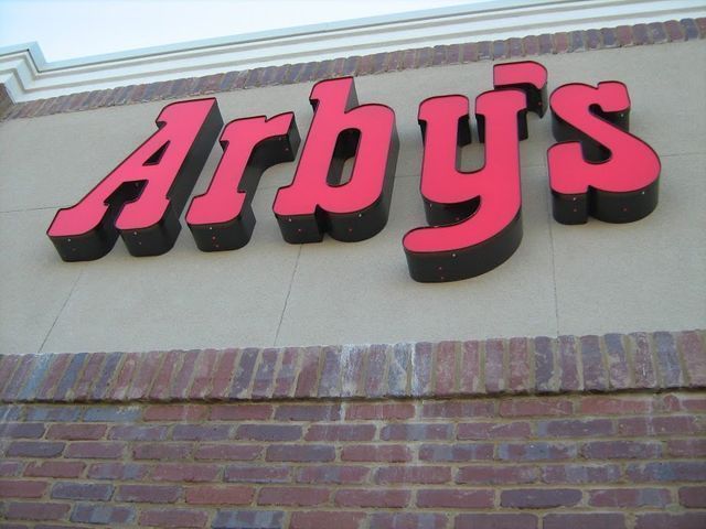 Arby's sign with red letters outlined in black, mounted on a tan building facade with brick base.