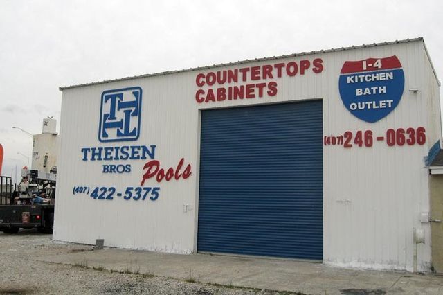 White building with blue roll-up door, signage for countertops, cabinets, pools, and a kitchen/bath outlet. Phone numbers are visible.