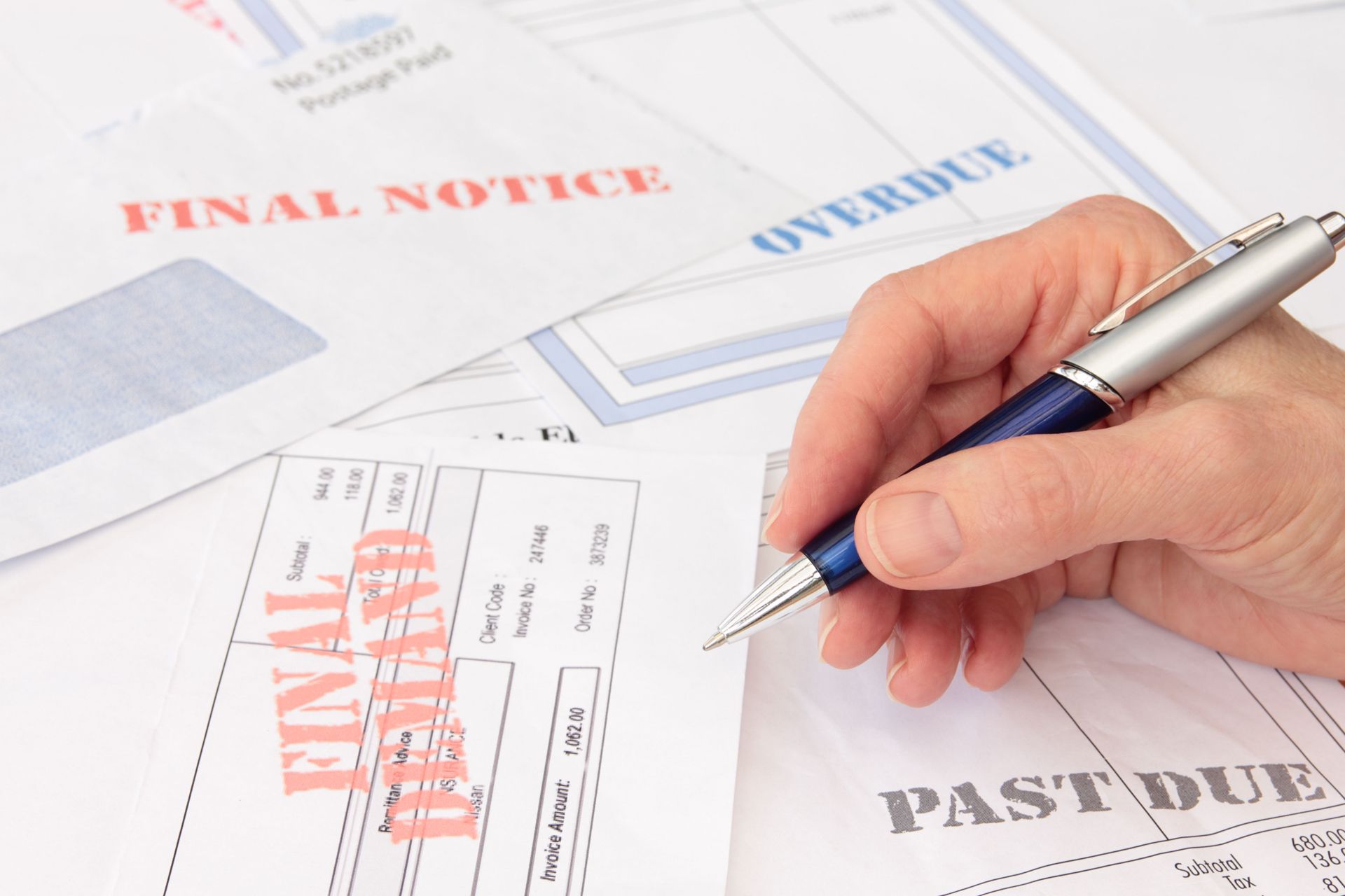 Hand holding a pen over overdue bills and final notices on a desk