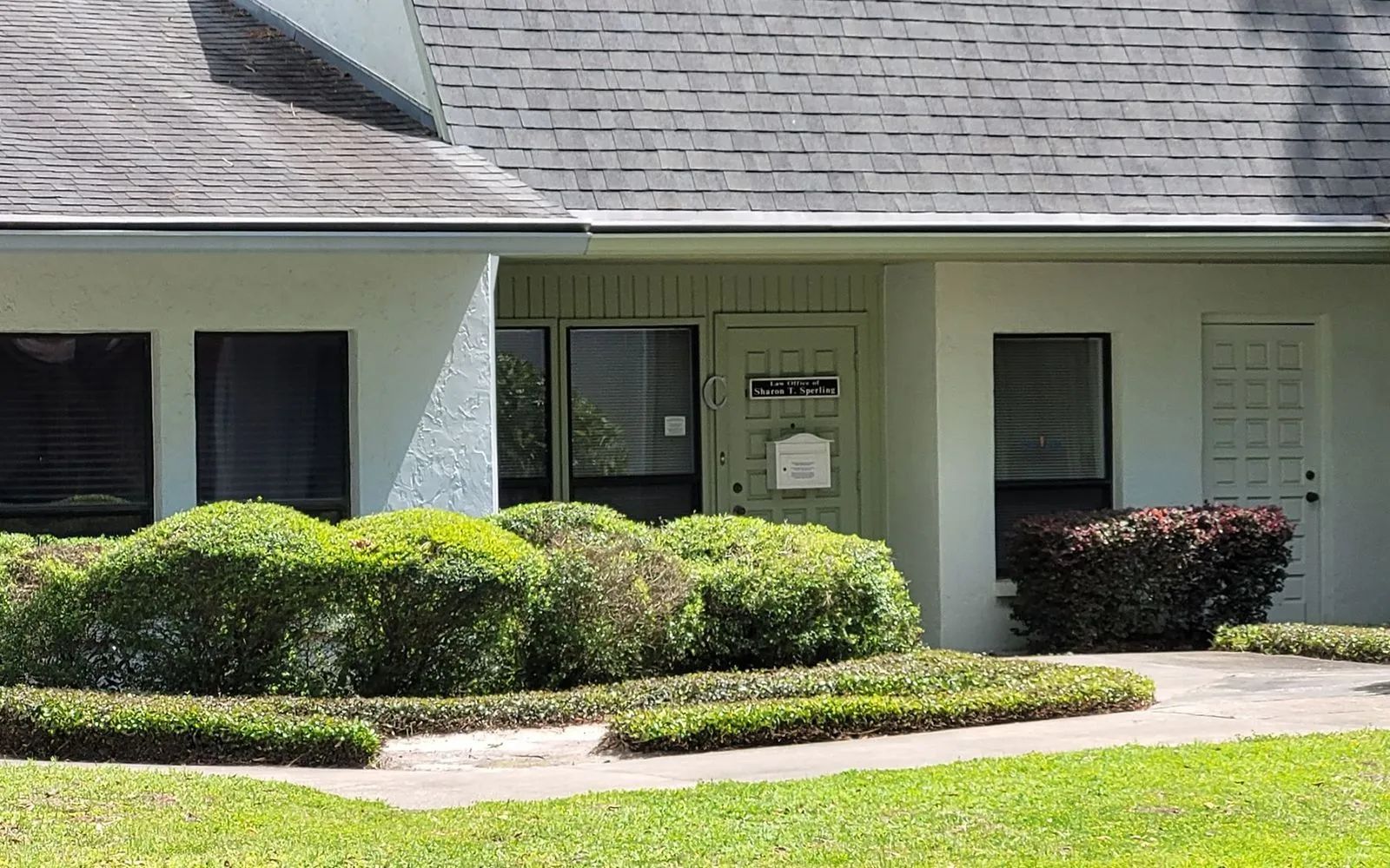Single-story house entrance with white siding, green shutters, shrubs, and a sidewalk in front