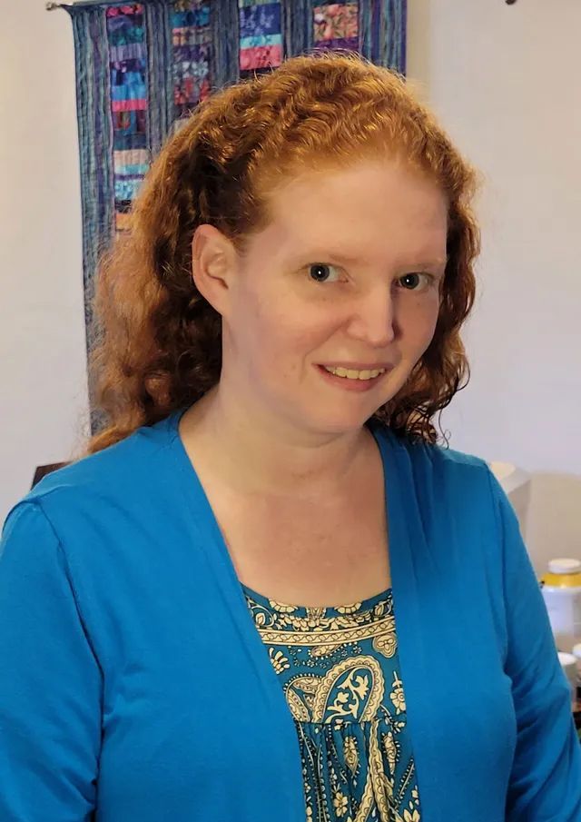 Smiling person in a blue cardigan with curly hair, standing indoors against a colorful wall hanging