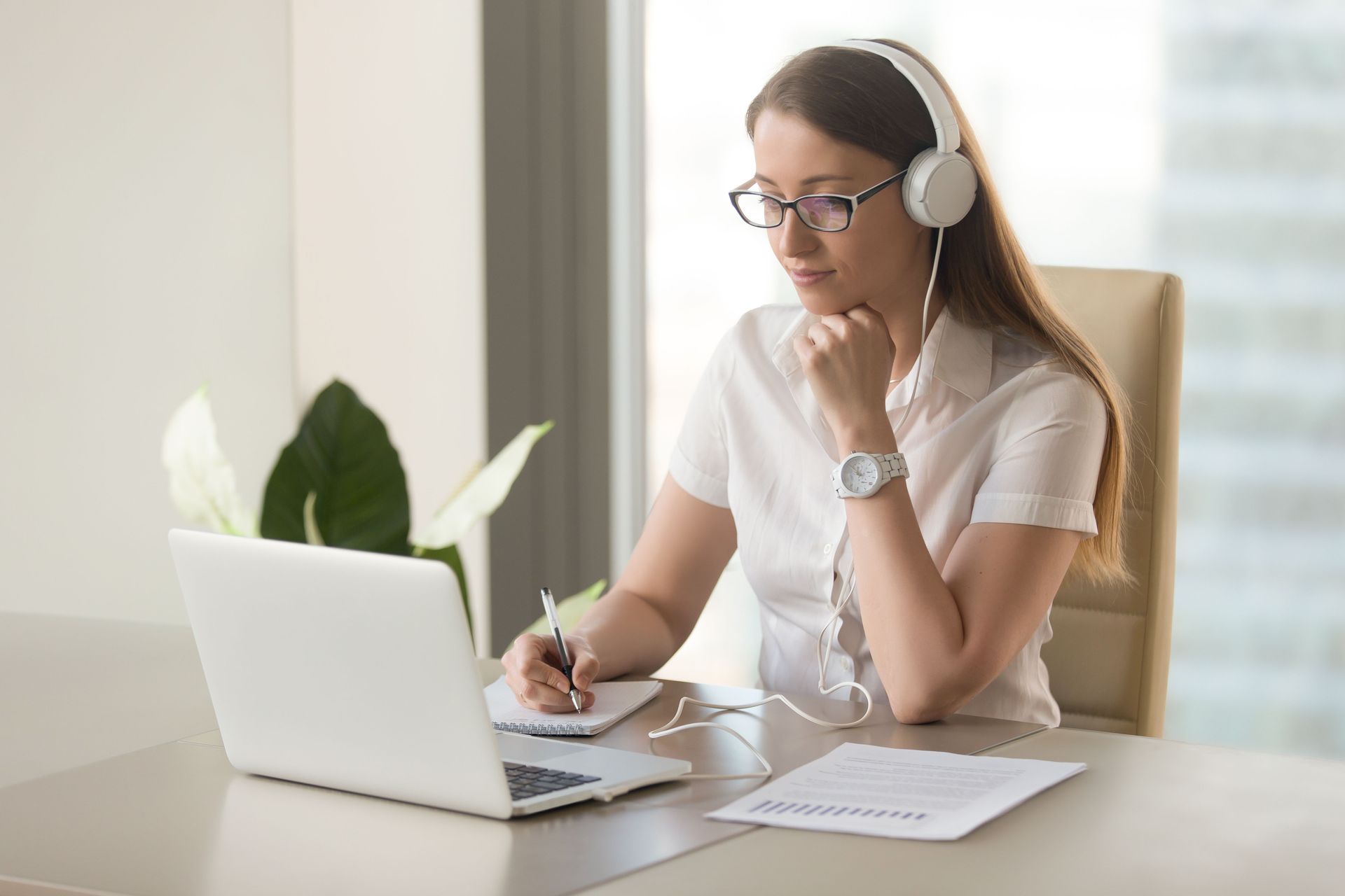 Woman working on a laptop at a desk, wearing headphones and writing notes in a bright office