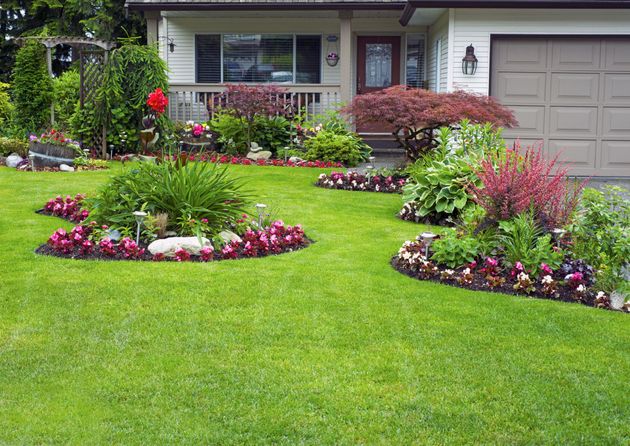 A landscaped front yard featuring lush green grass, curved flower beds with pink and red blooms, and ornamental shrubs.
