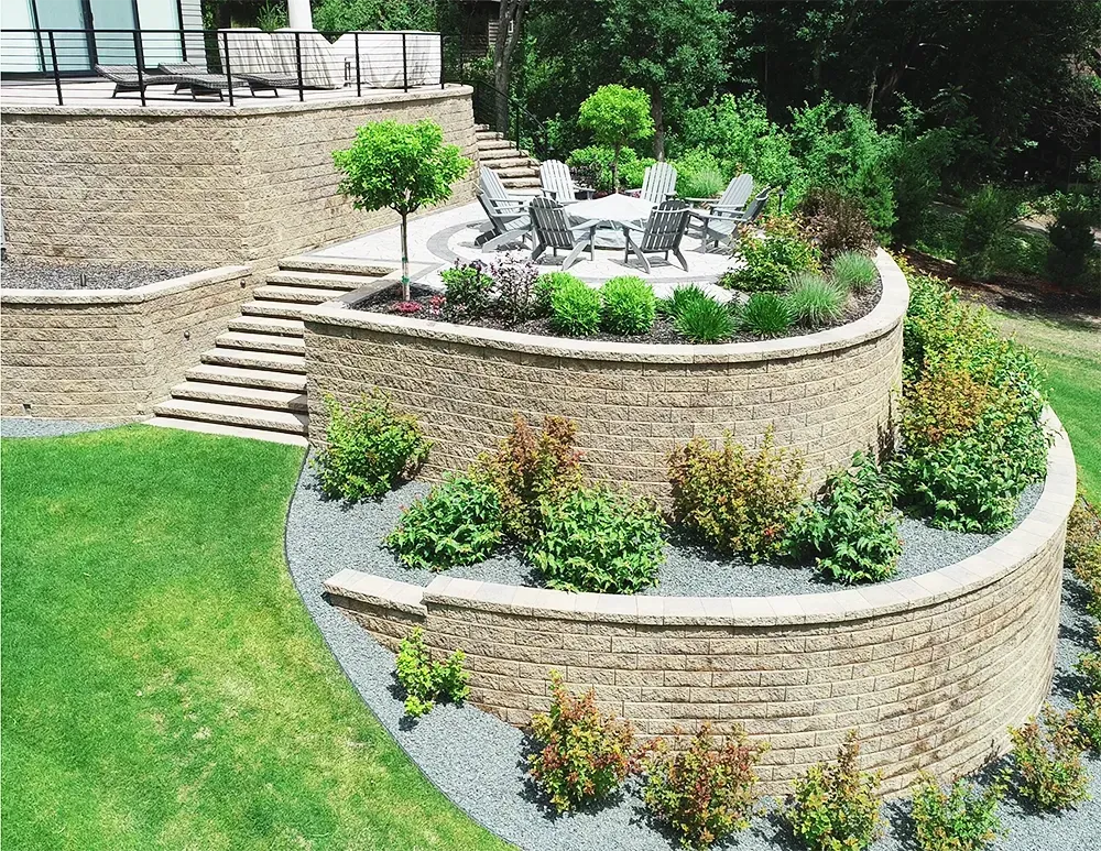 A multi-tiered stone retaining wall landscape featuring a patio with outdoor seating and lush shrubs, seen from above.