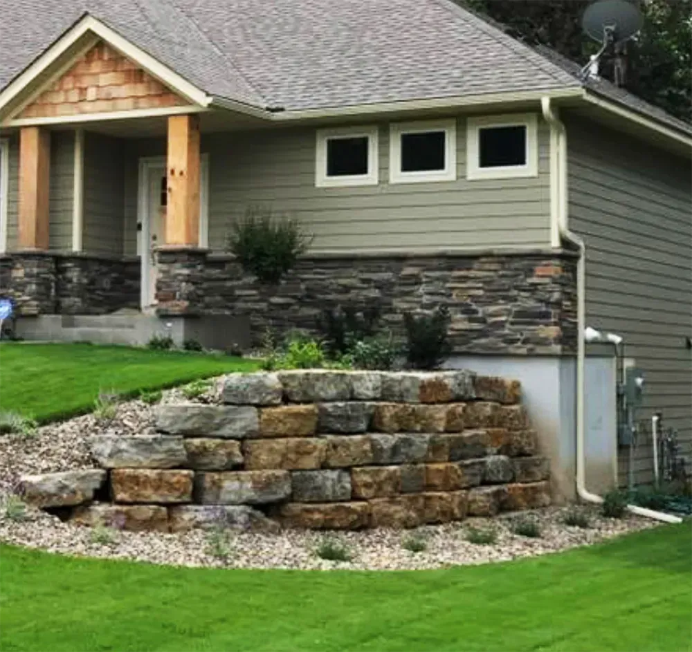 A tan-sided house with stone accents, featuring a large, tiered stone retaining wall set in a landscaped yard.