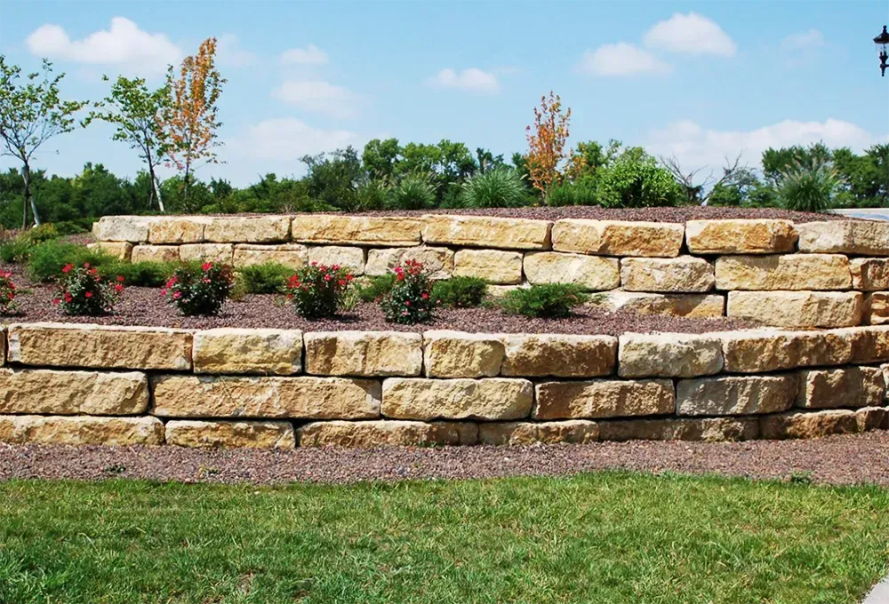 A two-tiered retaining wall made of stacked tan limestone blocks, surrounded by mulch, small shrubs, and a grassy lawn.