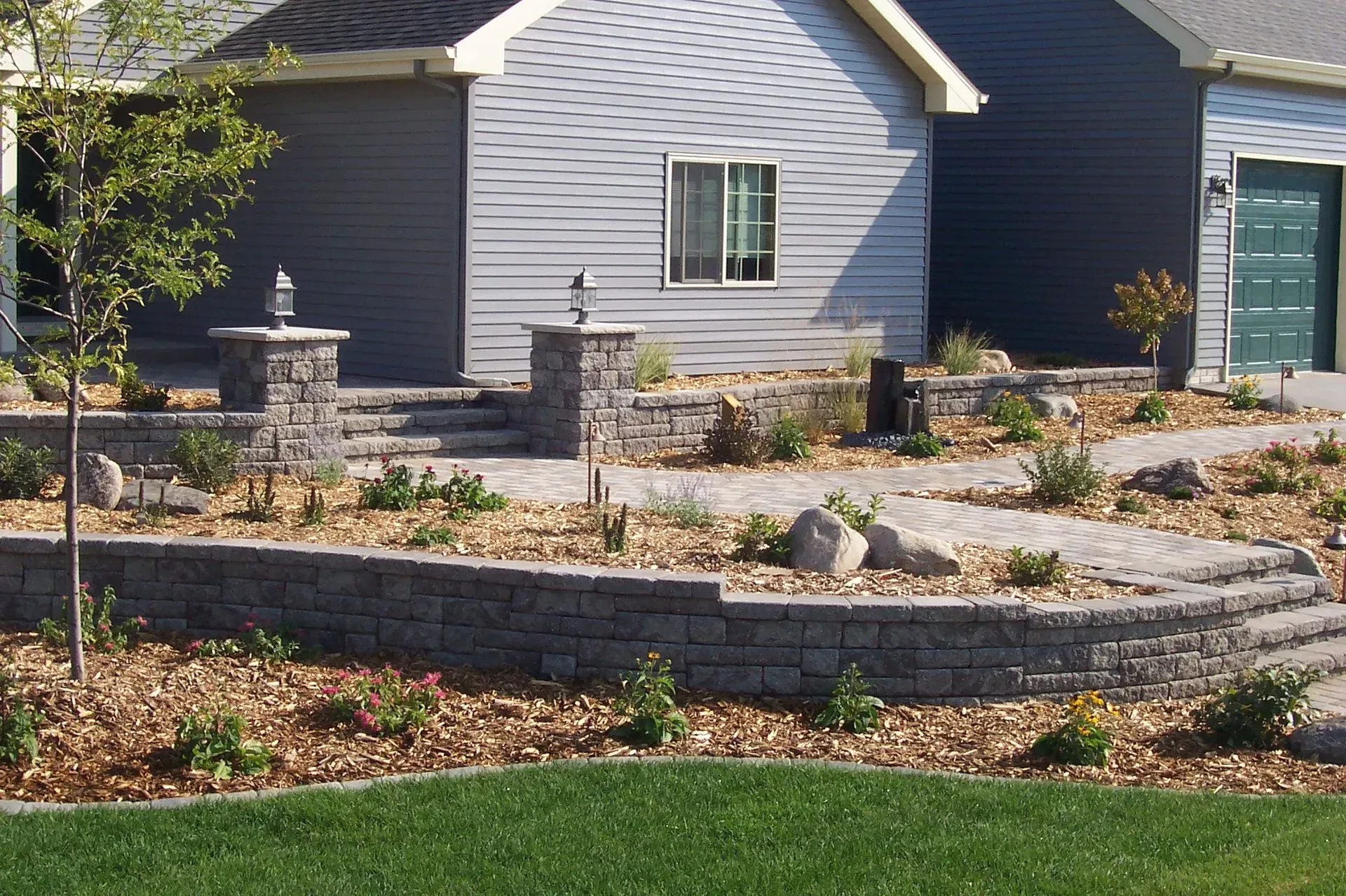 A multi-level stone retaining wall garden with paved pathways, landscape lighting, and small shrubs in front of a house.