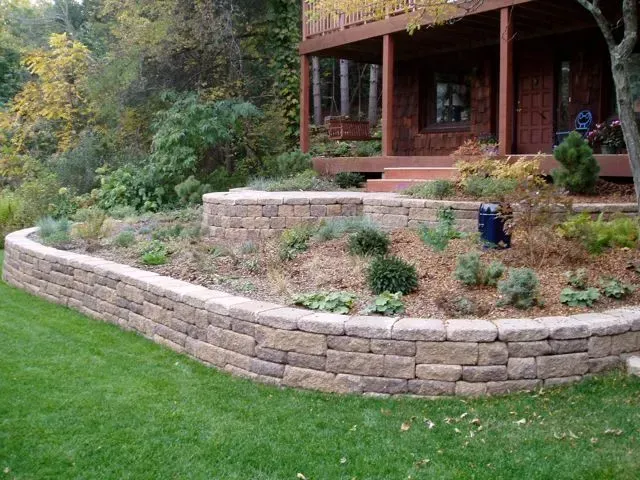 A multi-level stone retaining wall landscaped with various shrubs and plants in front of a rustic wooden deck.