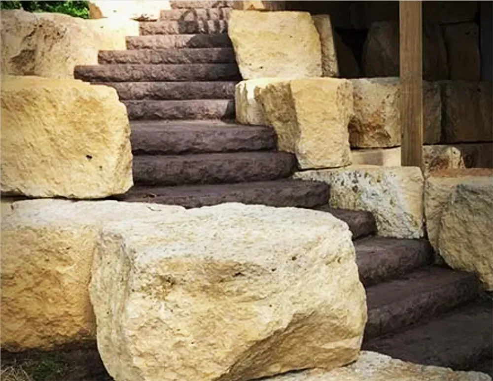 Outdoor stone stairs featuring dark treads flanked by large, tan, rough-hewn boulders.
