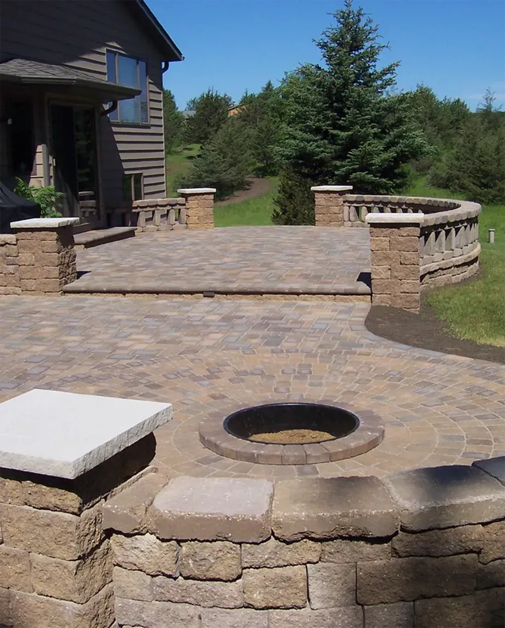 A tiered stone patio with a built-in circular fire pit, stone seating walls, and pillars outside a house.