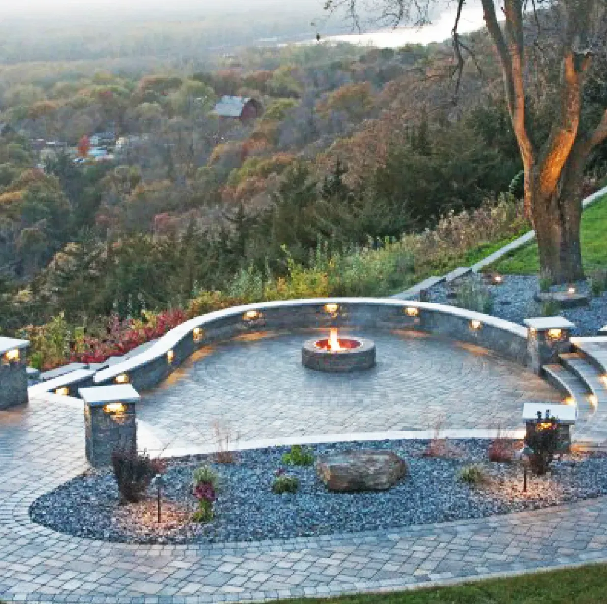 A paved outdoor patio with a circular fire pit, a stone retaining wall, and integrated lighting overlooking a forest.
