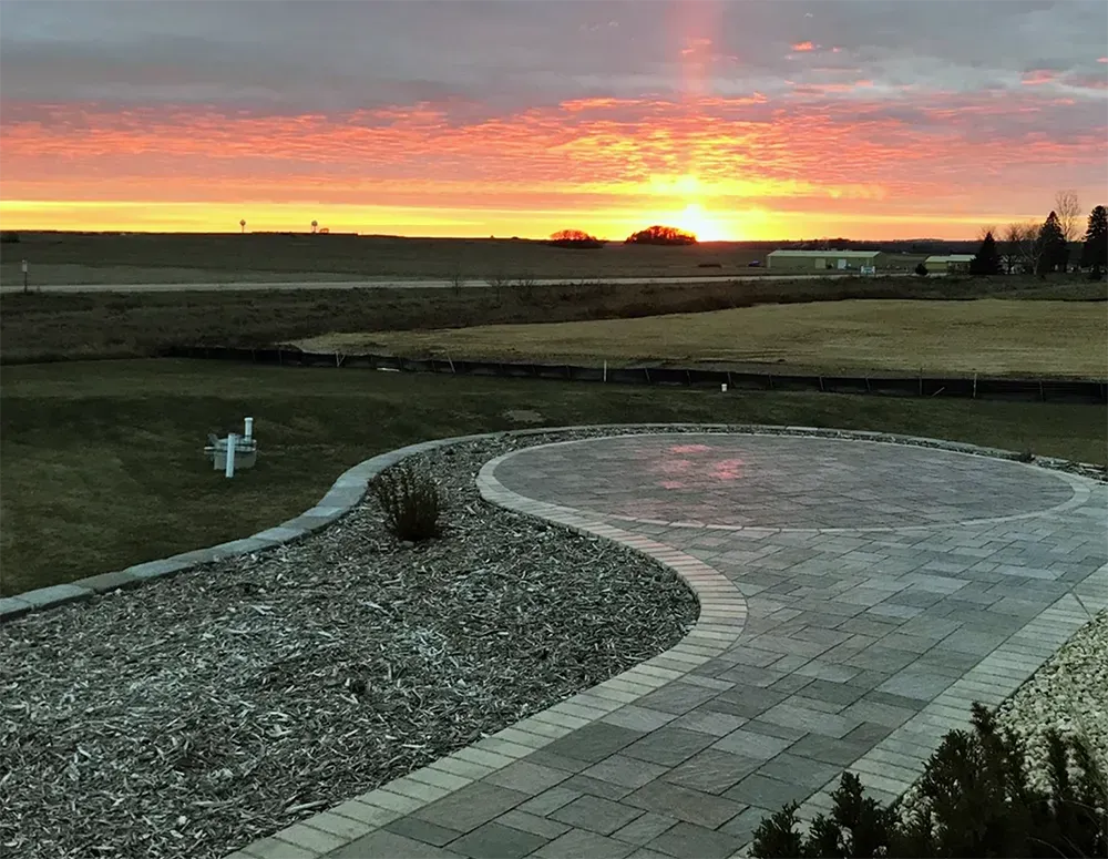 A stone walkway leads to a circular patio at sunset, overlooking a vast, grassy field under a bright orange sky.